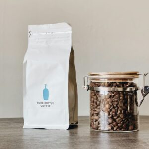 Bolsa blanca de café en grano junto a un frasco de vidrio lleno de granos tostados
