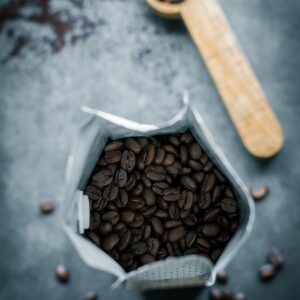 Bolsa abierta con granos de café tostado y una cuchara medidora de madera sobre una superficie azul oscuro