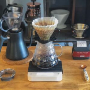 Preparación de café de filtro con dripper V60, jarra de vidrio, tetera y báscula sobre una mesa de madera